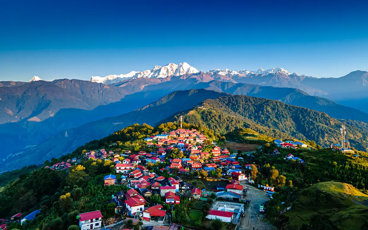 An aerial view shows a dense mountain village with red roofs nestled on a green ridge facing snowy peaks during the Annapurna Siklis Trek.