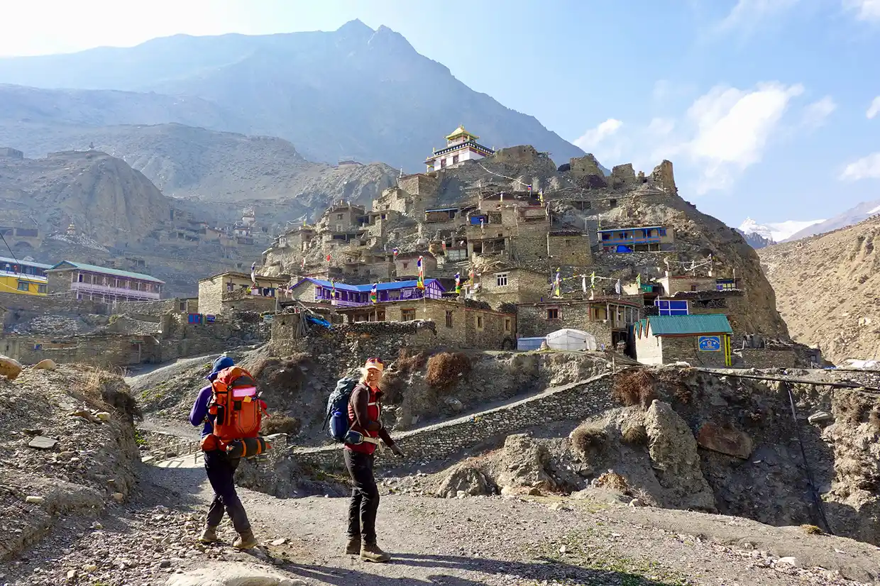 Two hikers with large backpacks walk on a dirt trail toward an ancient stone village built on a rocky hillside during the Nar Phu Valley with Annapurna Circuit Trek.