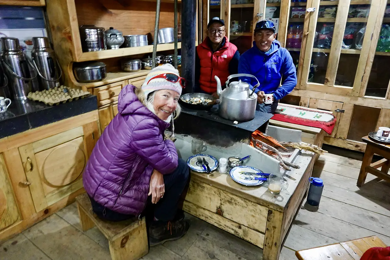 A smiling female trekker sits near a warm stove inside a traditional wooden teahouse kitchen during the Nar Phu Valley with Annapurna Circuit Trek.