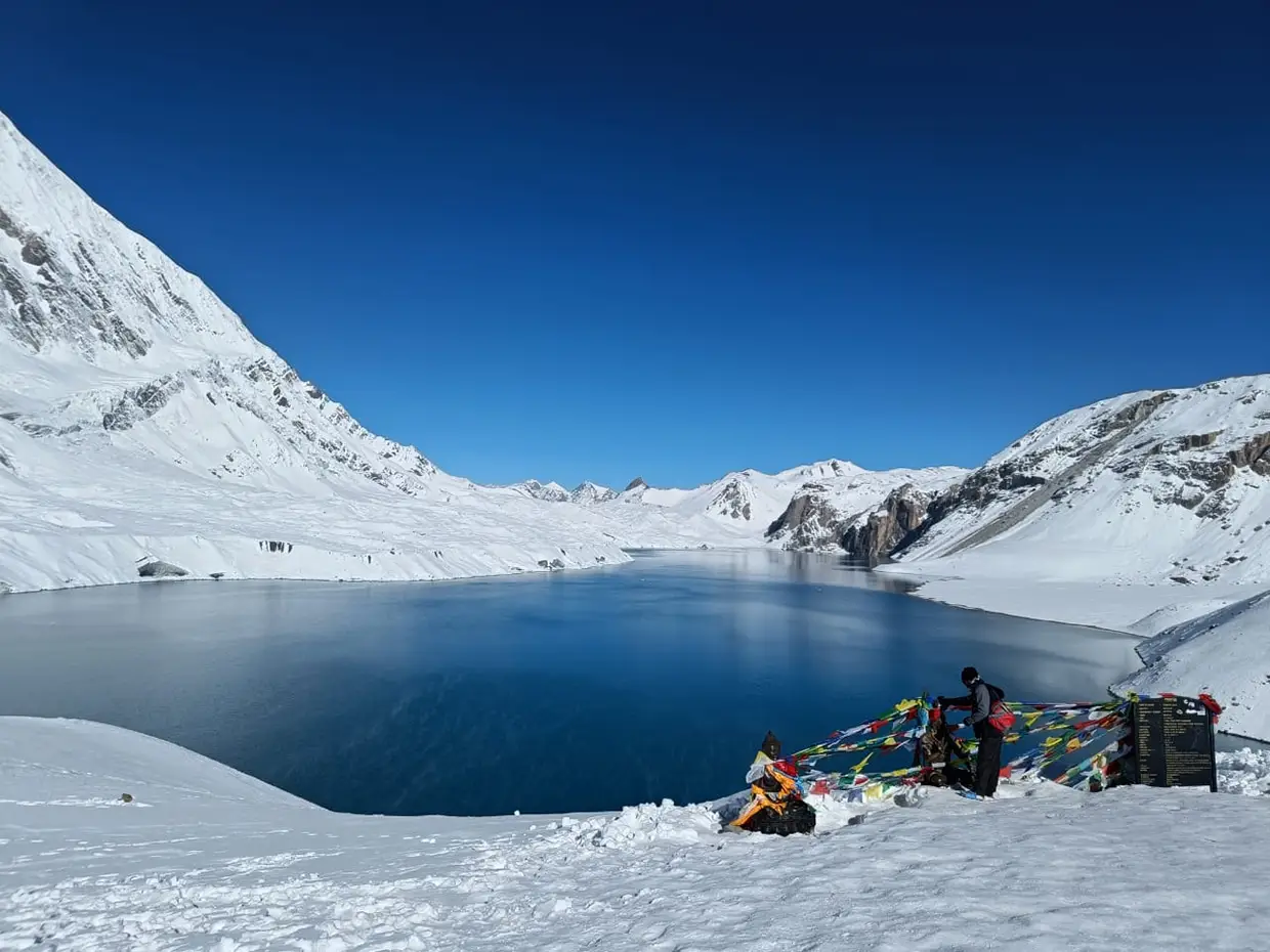 A clear blue lake is surrounded by snow-covered mountains and snowy ground, with hikers and prayer flags visible, during an Annapurna Circuit with Tilicho Lake Trek.