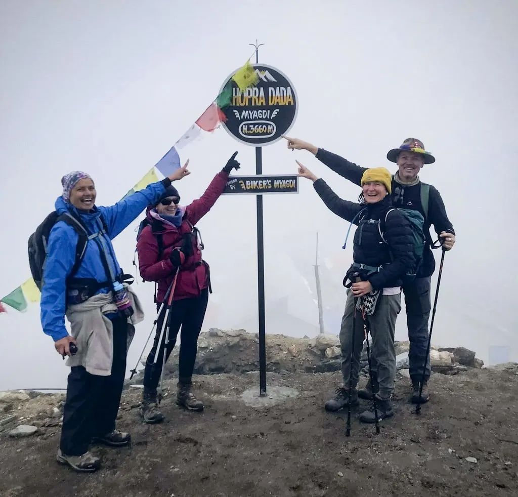Four smiling hikers celebrate reaching the Khopra Dada summit sign at 3,660 meters with prayer flags during their Khopra Ridge Trek.