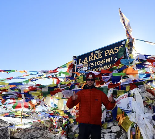 A hiker gives two thumbs up while standing in front of the Larke Pass sign at 5106m, heavily covered in colorful prayer flags during a trek in Nepal, perfect for a Manaslu Circuit Trek Guide.