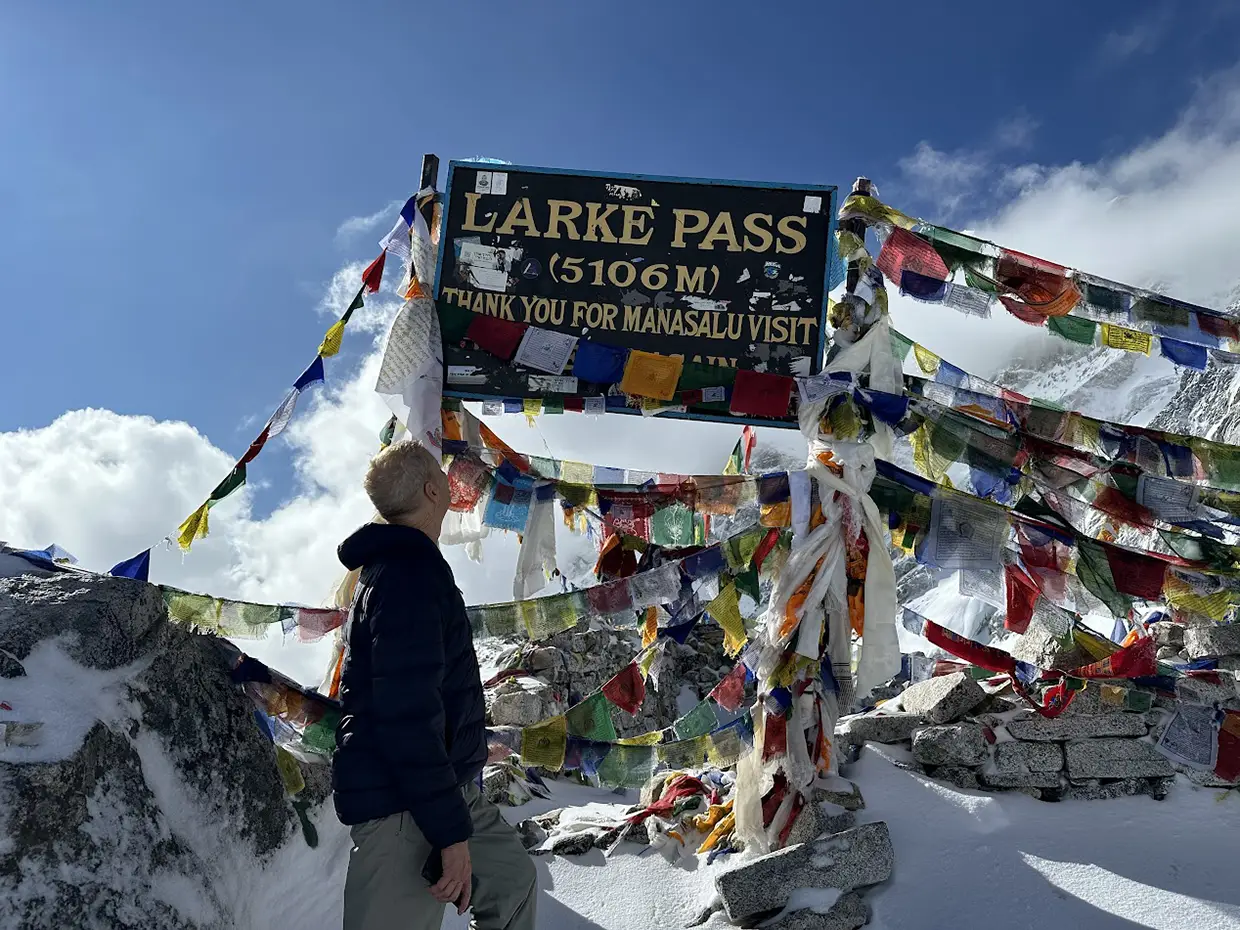 A trekker looks up at the Larke Pass sign surrounded by prayer flags and snow. Comparing the Manaslu vs Annapurna Circuit helps hikers prepare for high-altitude passes.