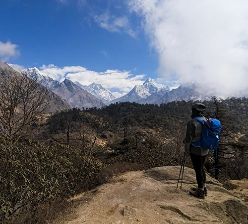 A hiker with a blue backpack stands on a rocky overlook, gazing at snow-capped Himalayan peaks. This Everest Panorama Trek Guide highlights spectacular mountain views along the route.