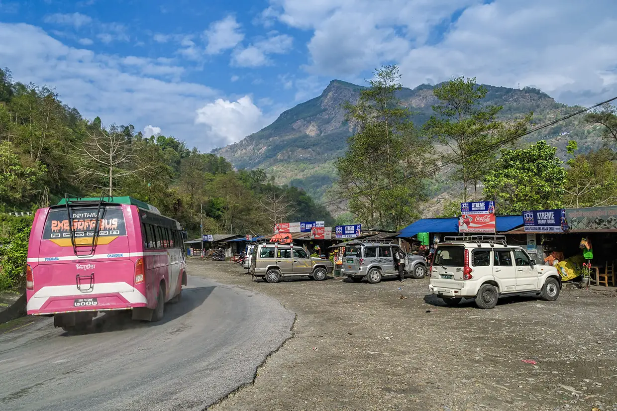A pink bus and several off-road vehicles park along a mountain road lined with shops, serving as a gateway for the Annapurna Community Trek.