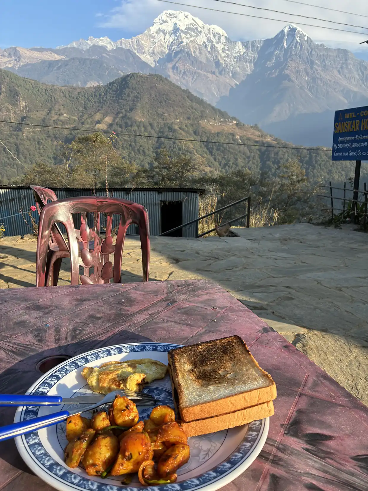 A plate of breakfast with toast, eggs, and potatoes sits on a table overlooking massive snow peaks, a highlight mentioned in this Poon Hill Trek Guide.