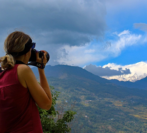 A woman in a magenta top uses a professional camera to photograph a snow-capped mountain peak and lush green valleys during the Annapurna Siklis Trek.
