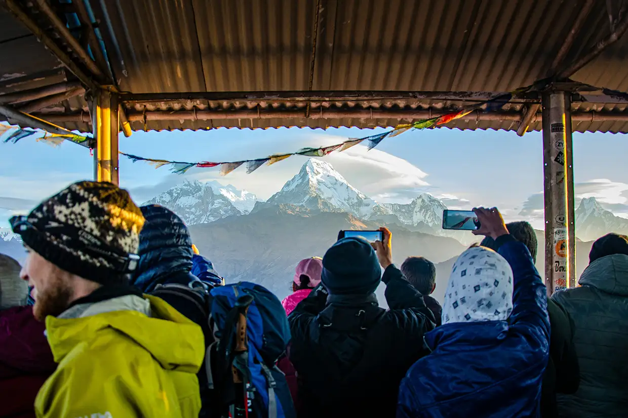 A crowd of trekkers uses smartphones to photograph a snow-capped mountain under a sheltered viewing platform. The Annapurna Panorama Trek features this bustling morning viewpoint.