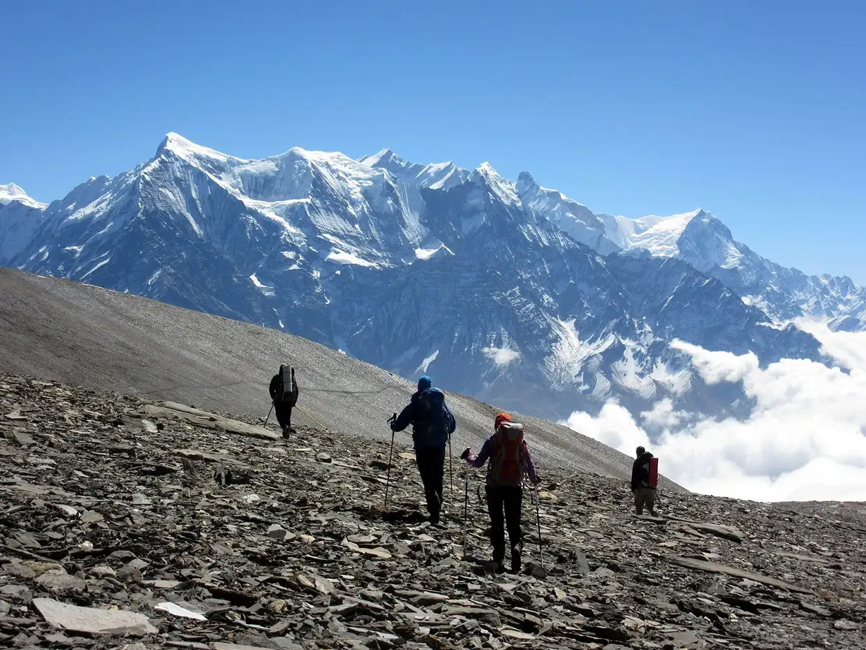 Trekkers with backpacks hike across a rocky mountain slope toward massive snow-capped peaks. This high-altitude scenery is a highlight of the Dhaulagiri Circuit Trek Guide.