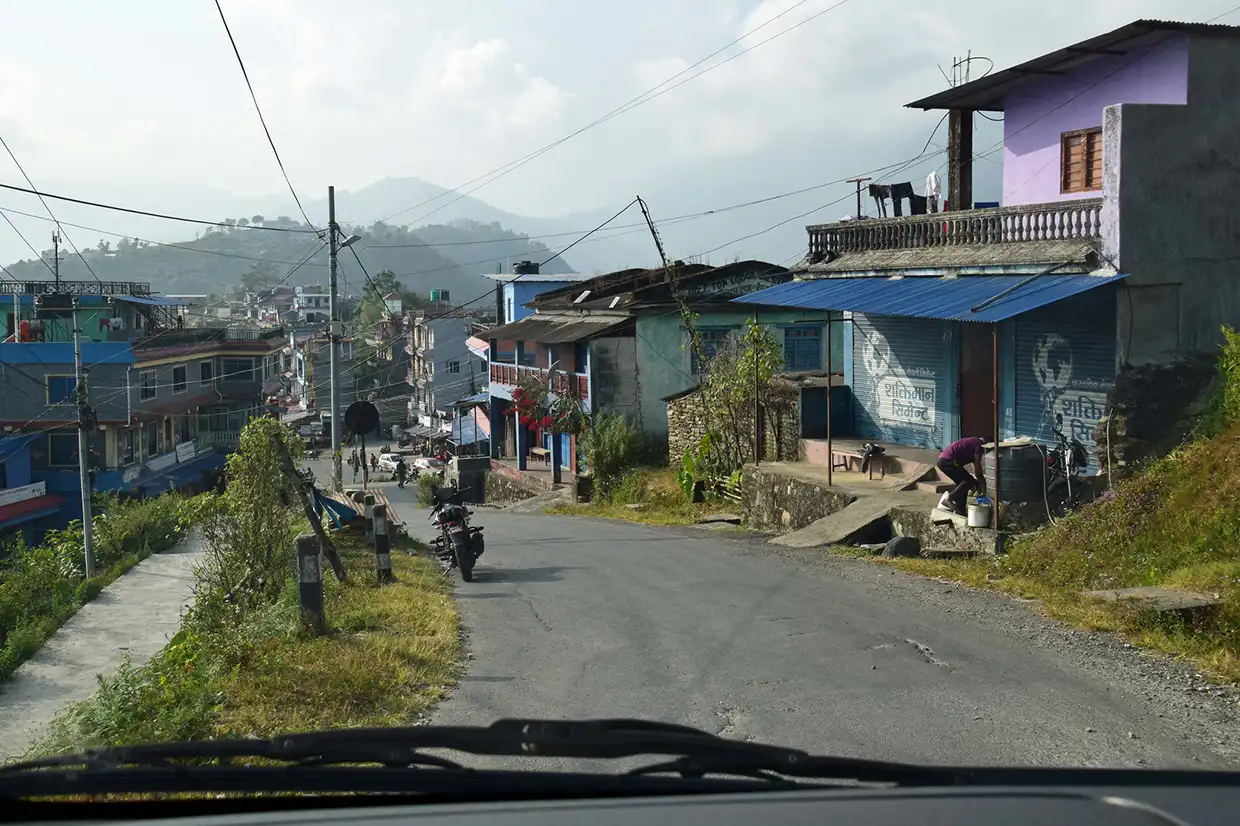 A winding paved road passes through a mountain town with buildings and hillsides. Travelers on the Annapurna Panorama Trek often drive through here.