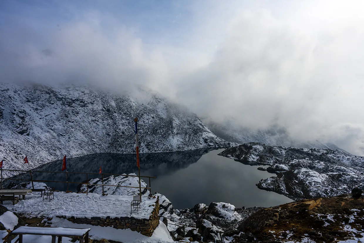 A tranquil, snow-dusted shoreline overlooks the dark waters of Gosaikunda Lake under misty skies, a serene destination often included when planning hiking in Nepal by month.
