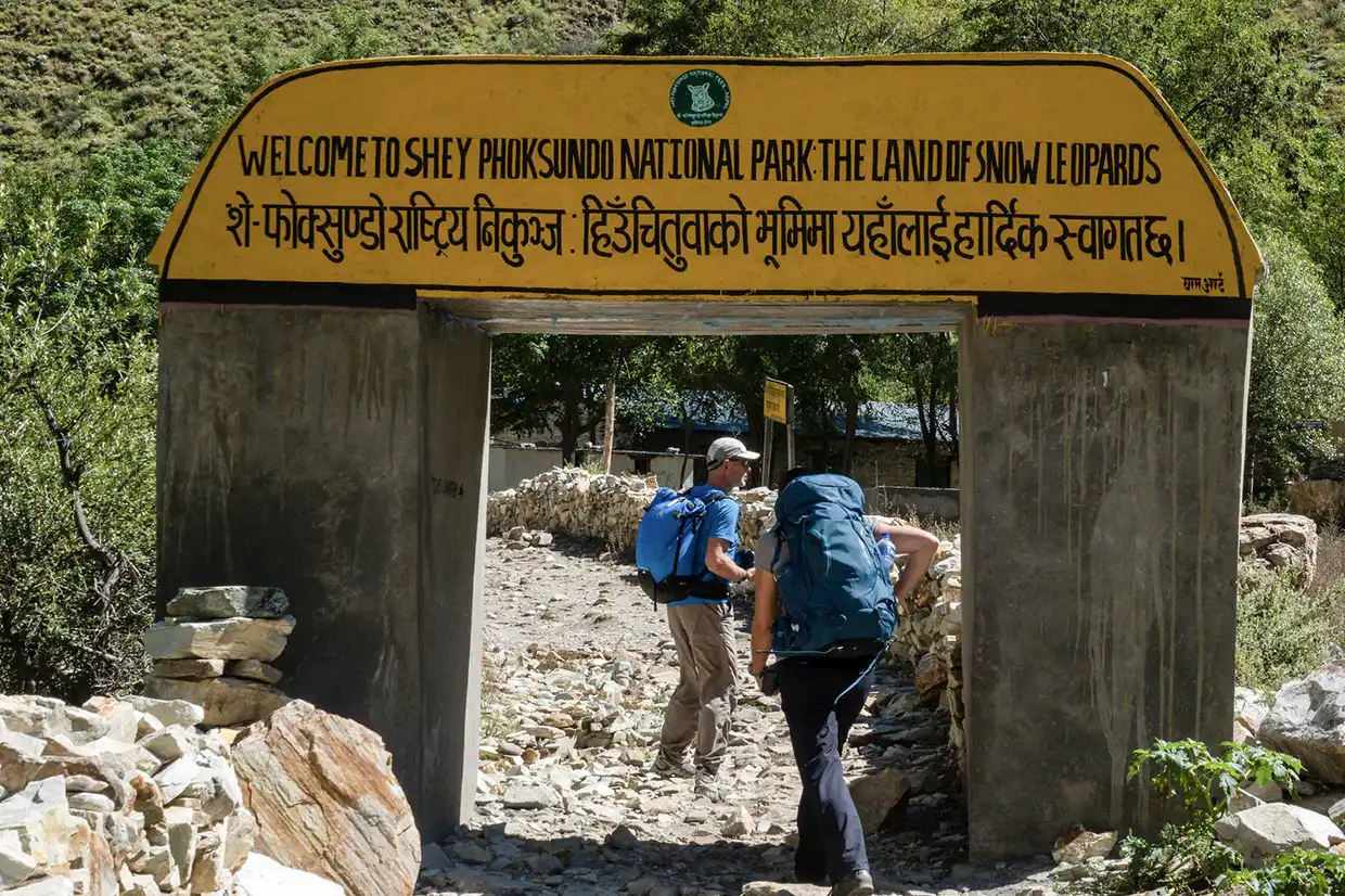 Two hikers with backpacks walk toward a yellow sign welcoming them to Shey Phoksundo National Park. Understanding current Nepal Trekking Regulations is essential for entering this remote, protected region.
