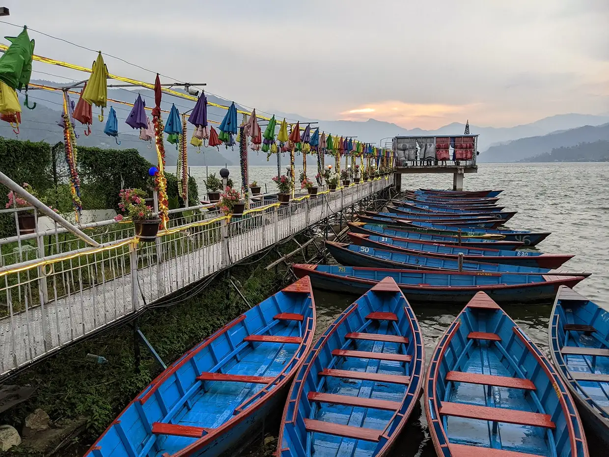 Colorful wooden boats are tied up along a decorative dock at sunset on Phewa Lake near Pokhara, a starting point for the Annapurna Community Trek.