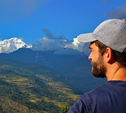 A male hiker wearing a grey cap looks out over terraced green hills toward a snow-capped mountain peak during the Annapurna Community Trek.