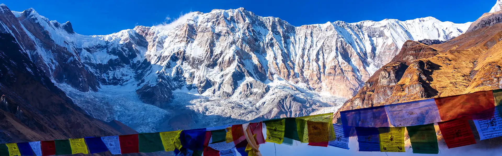 Colorful Buddhist prayer flags hang before a massive, snow-covered mountain wall and a sprawling white glacier, marking the path for a Thorong La Pass Crossing.