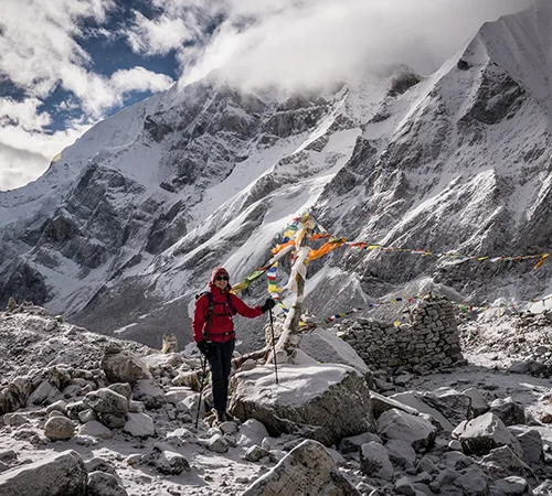 A trekker in a red jacket stands among snowy rocks and prayer flags beneath a massive Himalayan peak. Proper Fitness Training for Everest Base Camp trek helps travelers manage high-altitude conditions.