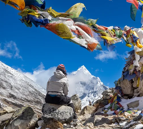A trekker sits on a rock below colorful prayer flags, looking at a towering snow peak under a bright blue sky on the Everest Cultural Trek.