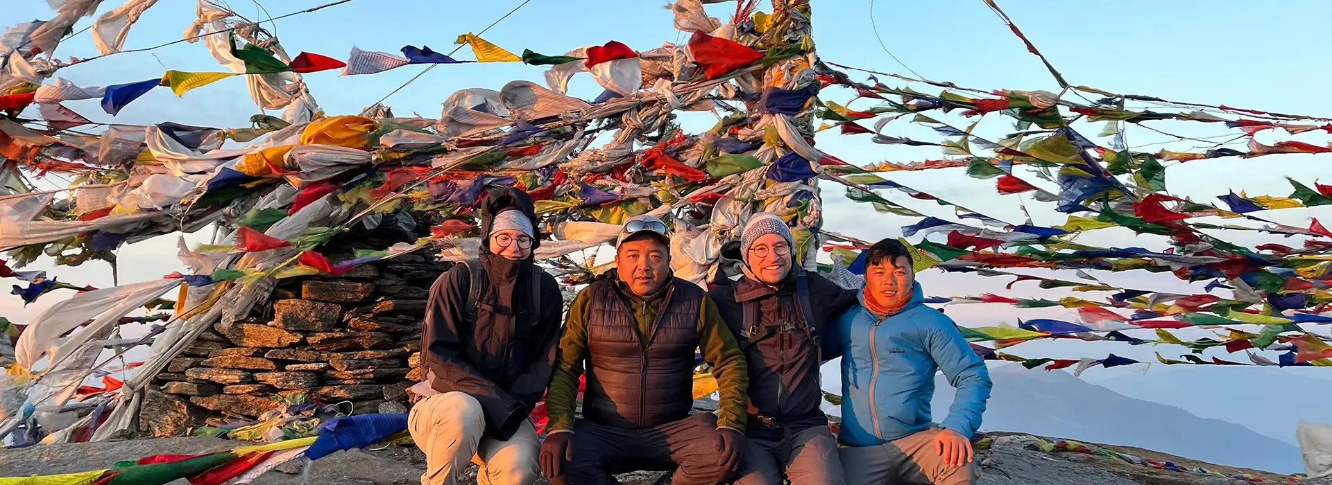 Four hikers sit together and smile in front of a large stone cairn wrapped in colorful Buddhist prayer flags during a Pikey Peak Trek.