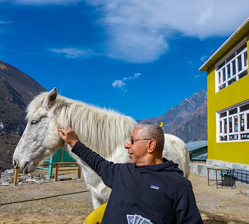 A traveler strokes a white horse next to a yellow building on a Tamang Heritage Trek, with rugged Himalayan peaks rising under a blue sky.