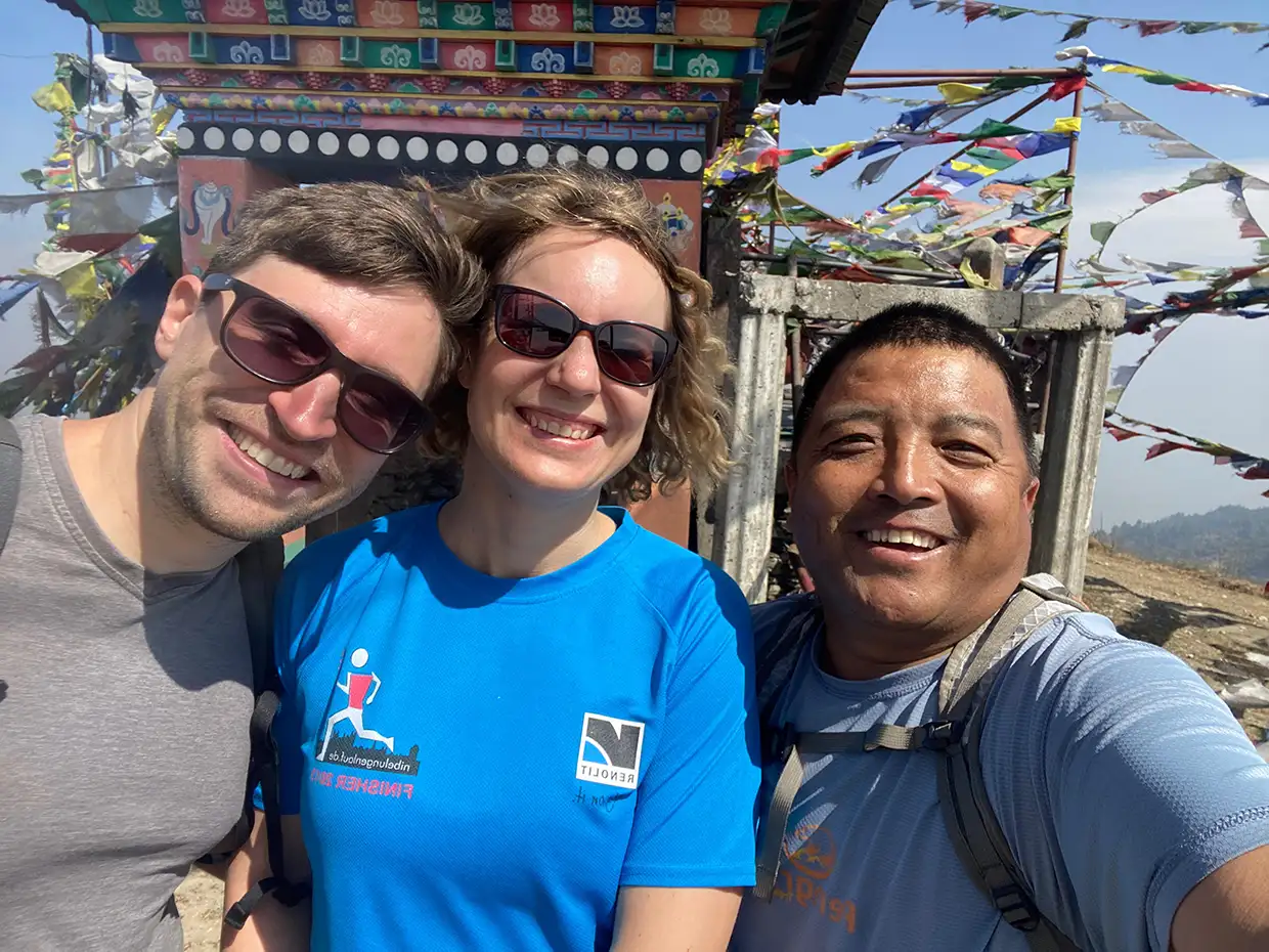Two travelers and their local guide smile for a close-up photo in front of a colorful Buddhist shrine decorated with prayer flags during a Pikey Peak Trek.