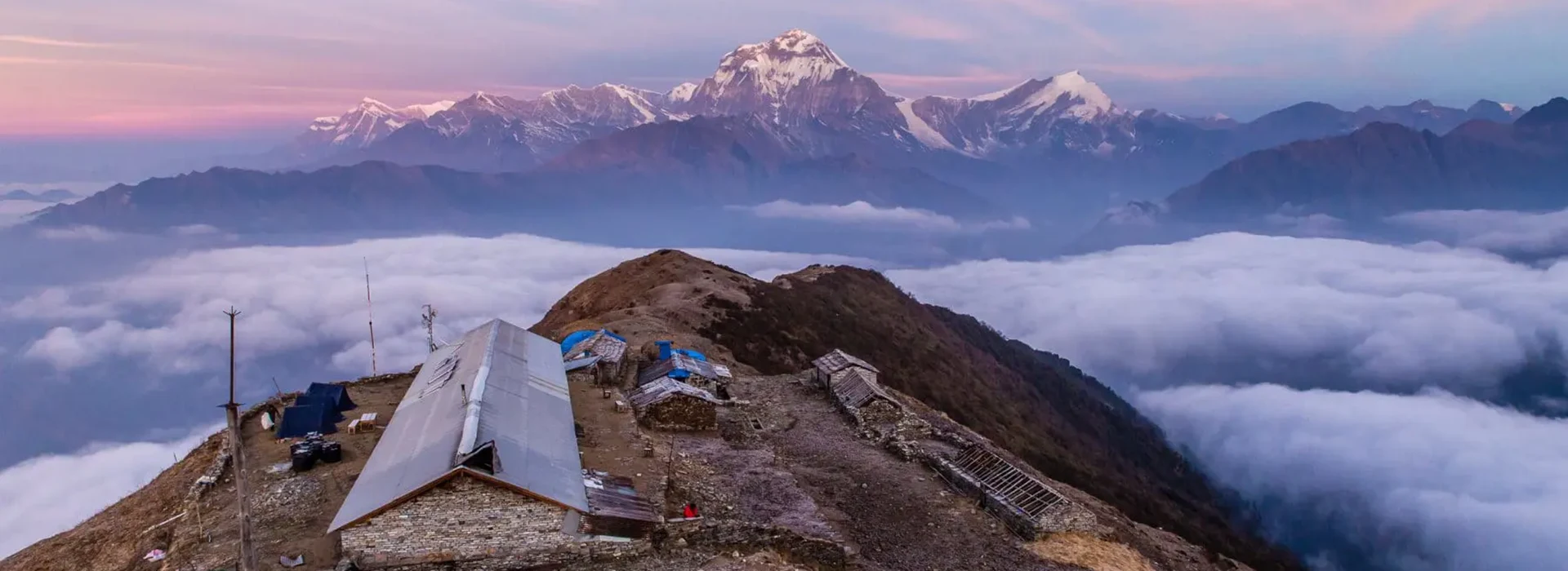 A mountain lodge sits on a grassy ridge above a blanket of clouds, facing snow-capped Himalayan peaks during a Khopra Ridge Trek sunrise.