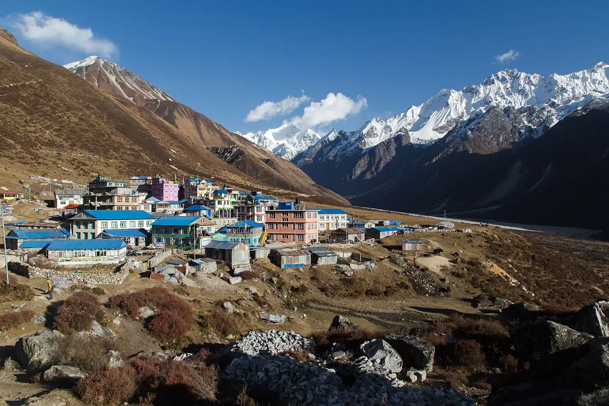 A colorful mountain village sits beneath towering snow-capped Himalayan peaks under a clear blue sky, illustrating a key destination found in our Langtang Valley Trek Guide.