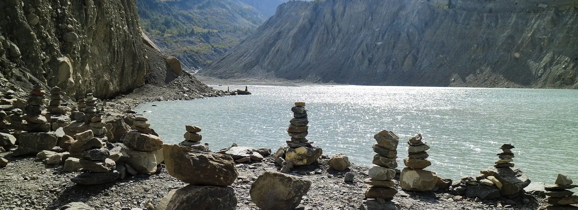 Several stone cairns stand on a rocky shore next to a shimmering glacial lake nestled within a deep valley on the Annapurna Circuit with Tilicho Lake Trek.