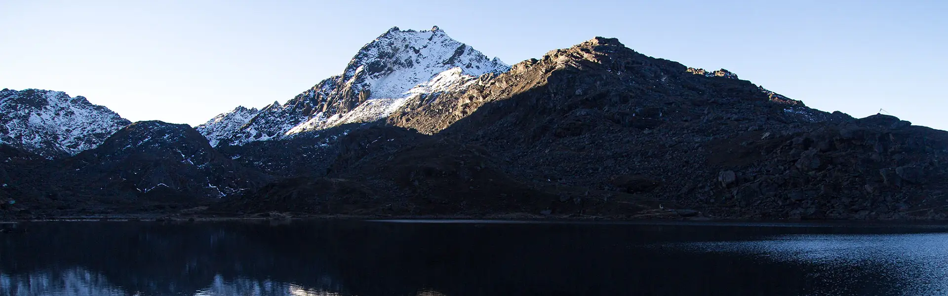 The morning sun illuminates a snowy Himalayan peak reflecting in the still, dark waters of a high-altitude lake along the Langtang Gosaikunda Trek Guide route.