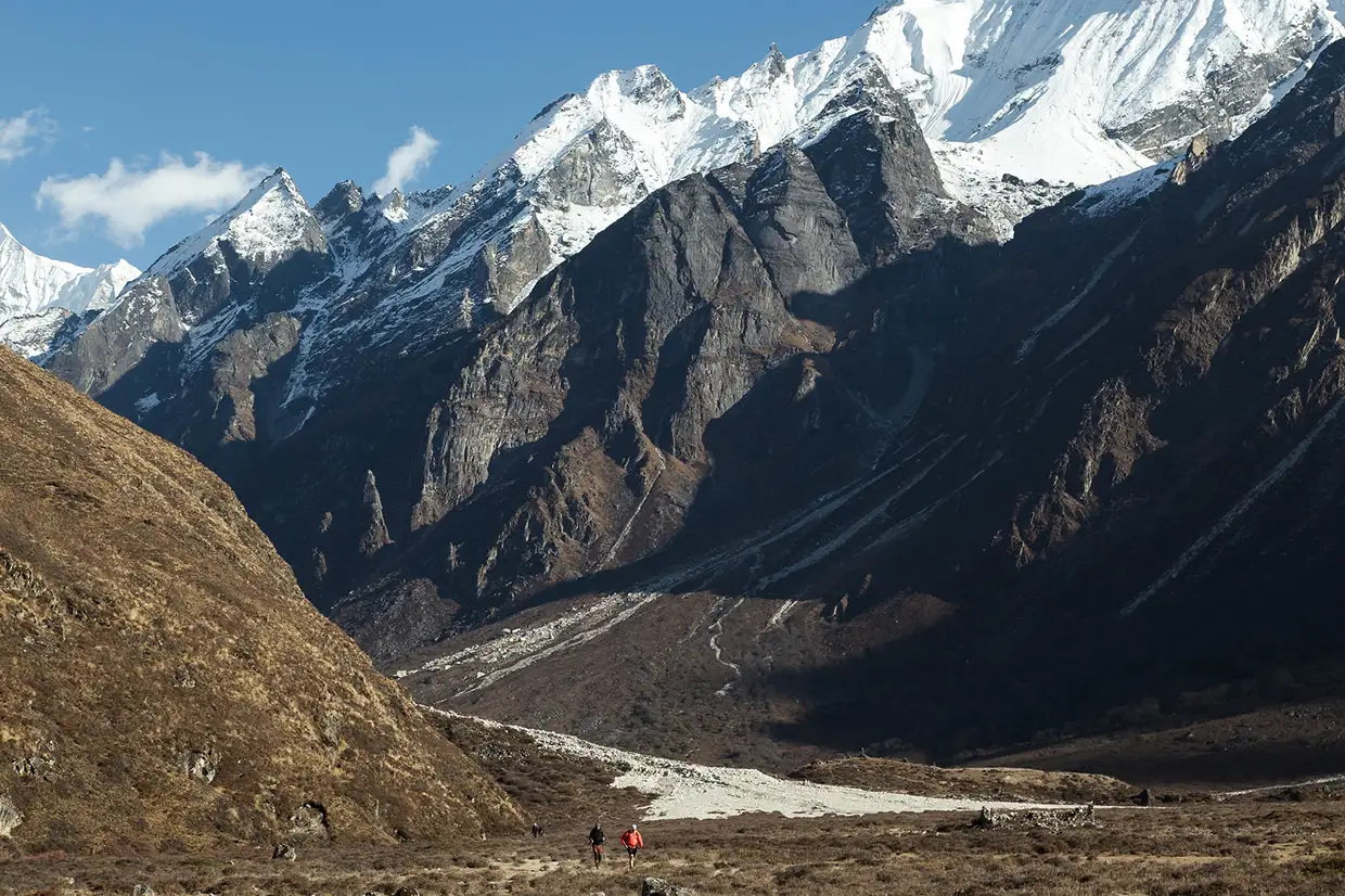 Two hikers walk through a wide mountain valley toward towering, jagged rock peaks and snow-covered summits. This rugged path is detailed in our Langtang Valley Trek Guide.