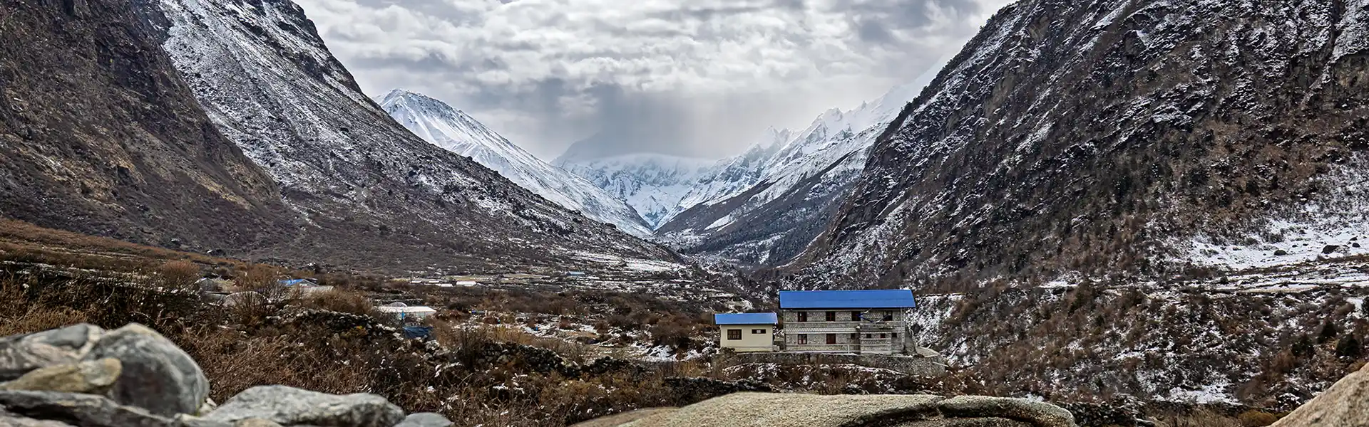 Small stone buildings with blue roofs sit nestled in a deep mountain valley surrounded by snow-covered peaks, an essential stop considered in any Langtang Trek Cost.