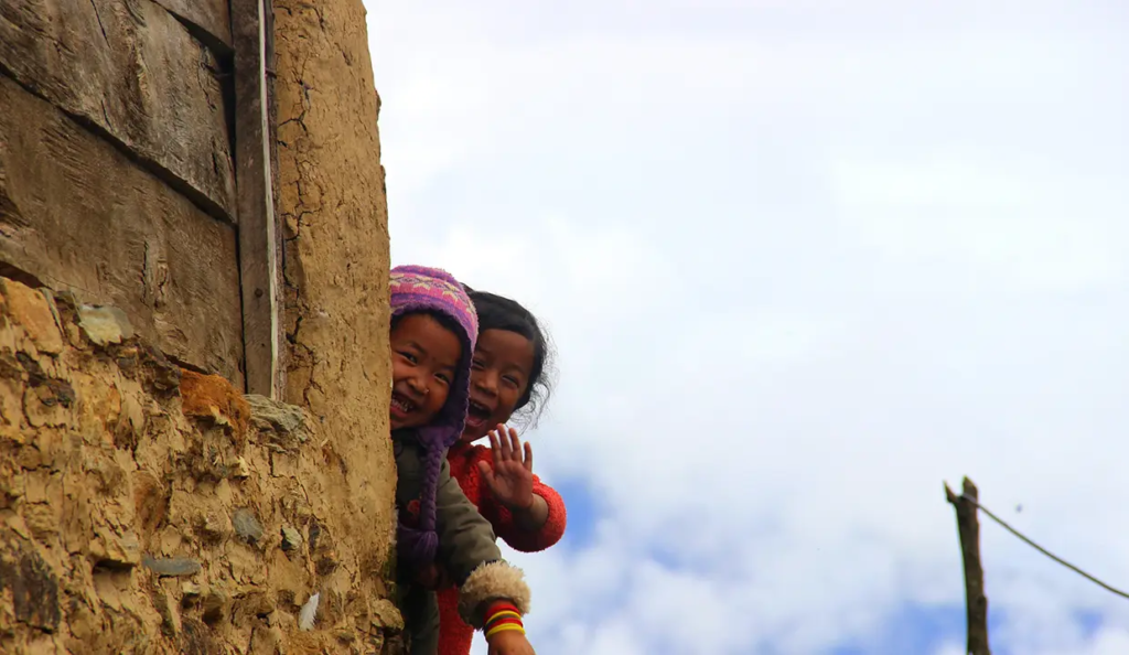 Two smiling young girls wave goodbye from behind a traditional mud brick house during the Annapurna Siklis Trek in Nepal.