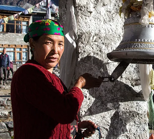 A local woman in a vibrant headscarf rings a traditional bell at Kyangjin Gonpa, a spiritual highlight found in any Langtang Valley Trek Guide.