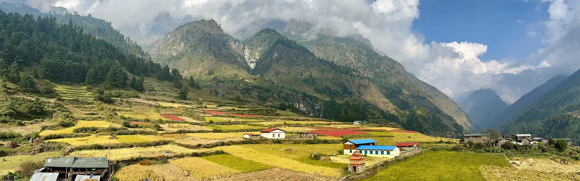 A wide panoramic view shows colorful agricultural fields and traditional village homes nestled under large, cloud-capped mountains during the Best Time for the Manaslu Circuit Trek.