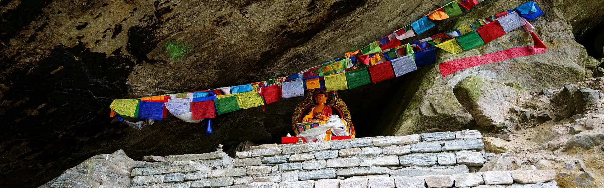 A golden Buddha statue rests within a rocky mountain cave adorned with vibrant prayer flags. This Annapurna Circuit Trek Guide highlights the unique cultural landmarks found along the trail.