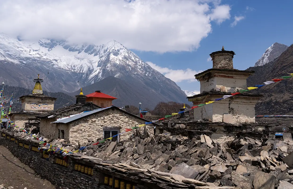 Ancient stone stupas and prayer wheels sit before a remote village and snowy peaks, showing the cultural wonders found while Hiking in Nepal by Month.