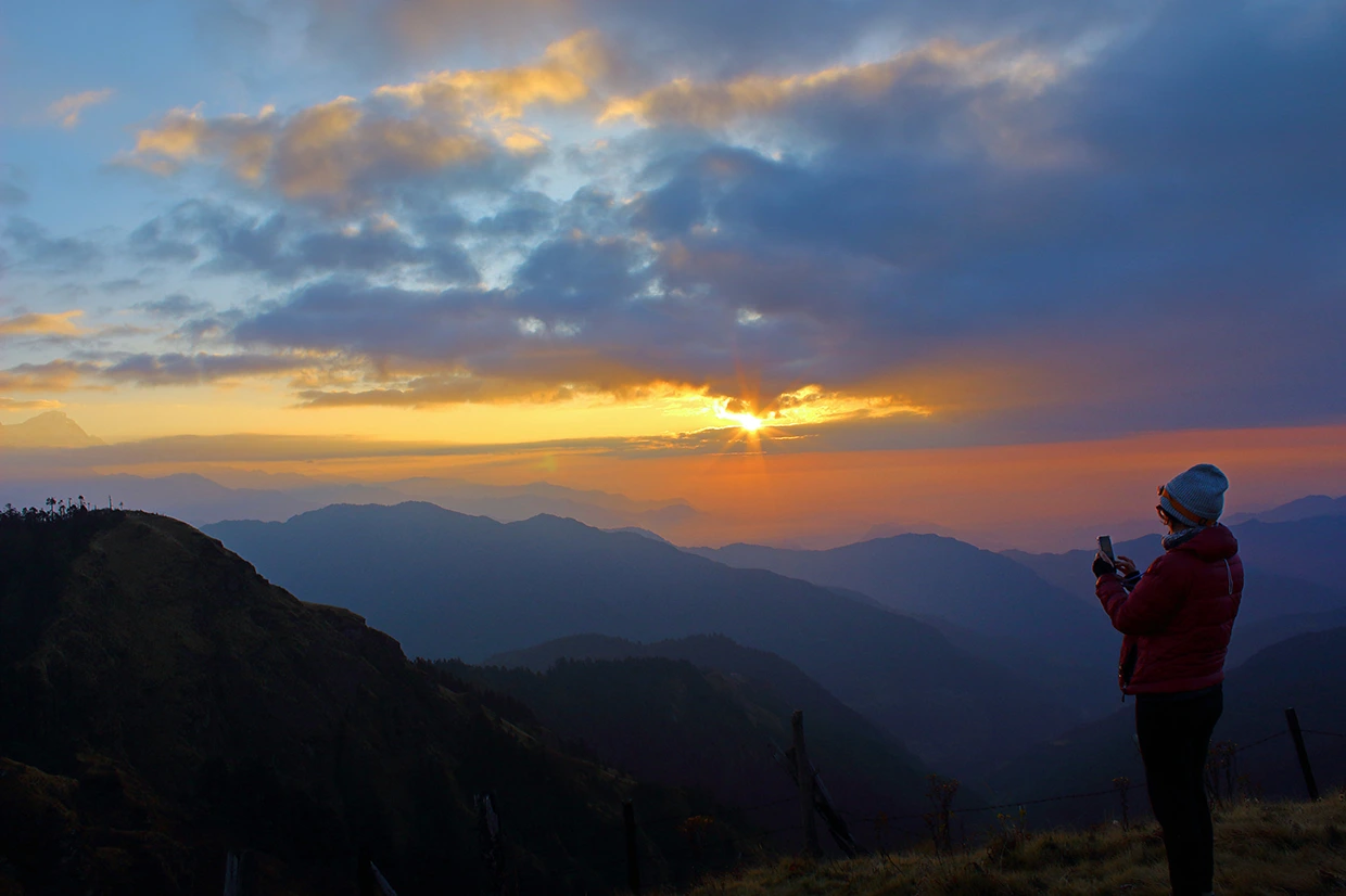 A lone hiker in a red jacket photographs a dramatic golden sunrise breaking through dark clouds over distant mountains during the Mohare Danda Trek.