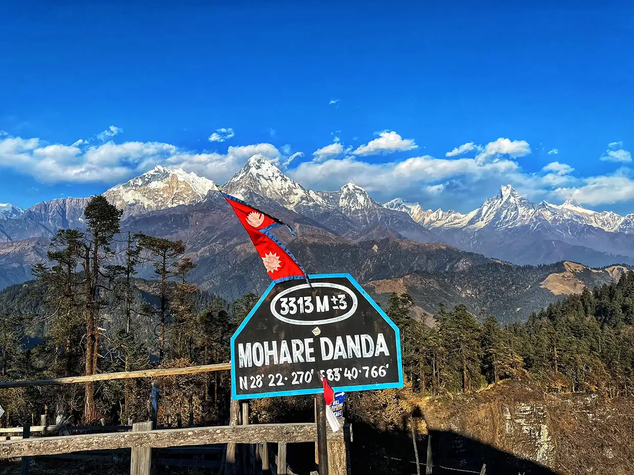 A wooden sign with "Mohare Danda Trek" coordinates sits on a viewpoint overlooking vast, snow-capped mountains under a bright blue sky with clouds.