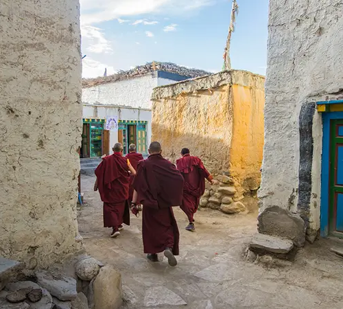 Four monks in red robes walk along a narrow stone pathway between traditional buildings in Upper Mustang, a region requiring Restricted Area Permits in Nepal.