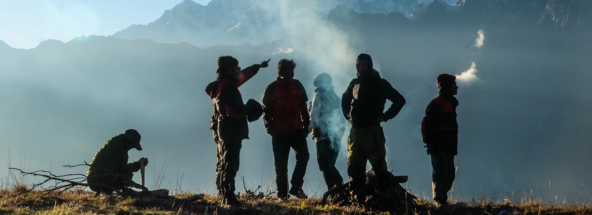 A group of hikers stands around a smoky campfire on a grassy ridge during a misty morning on the Annapurna Siklis Trek.