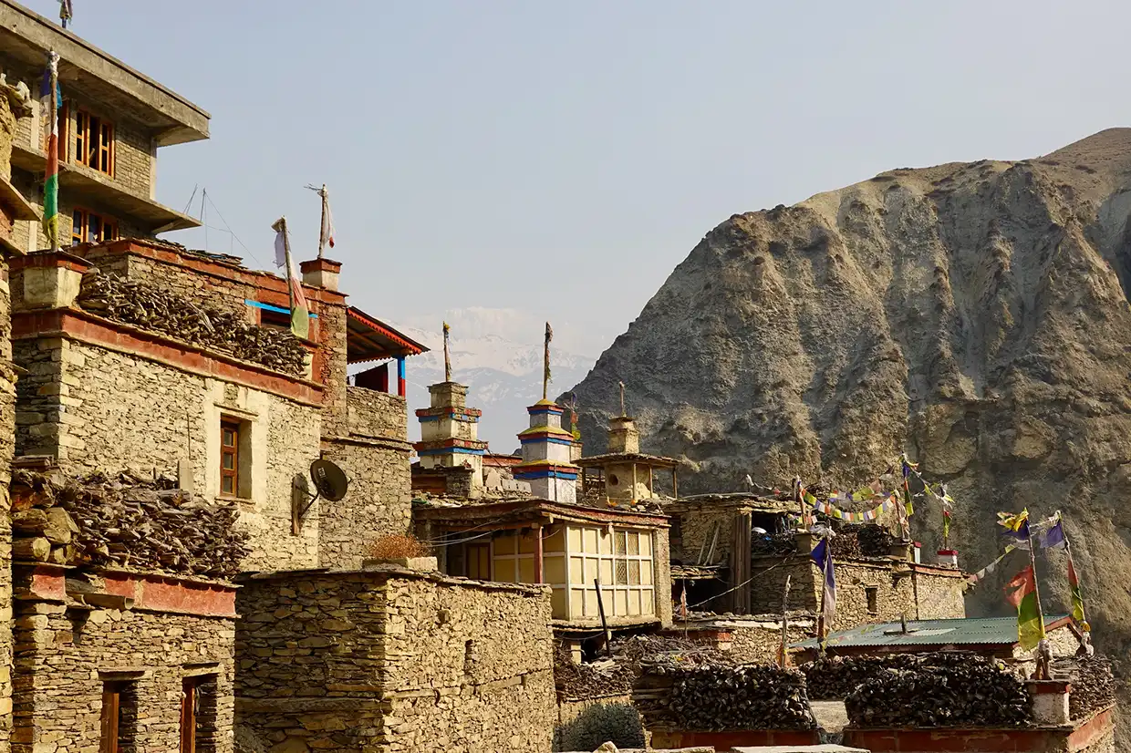 The stone-built traditional houses and chortens of Nar Phedi Monastery stand against a massive rocky cliffside during the Nar Phu Valley with Annapurna Circuit Trek.