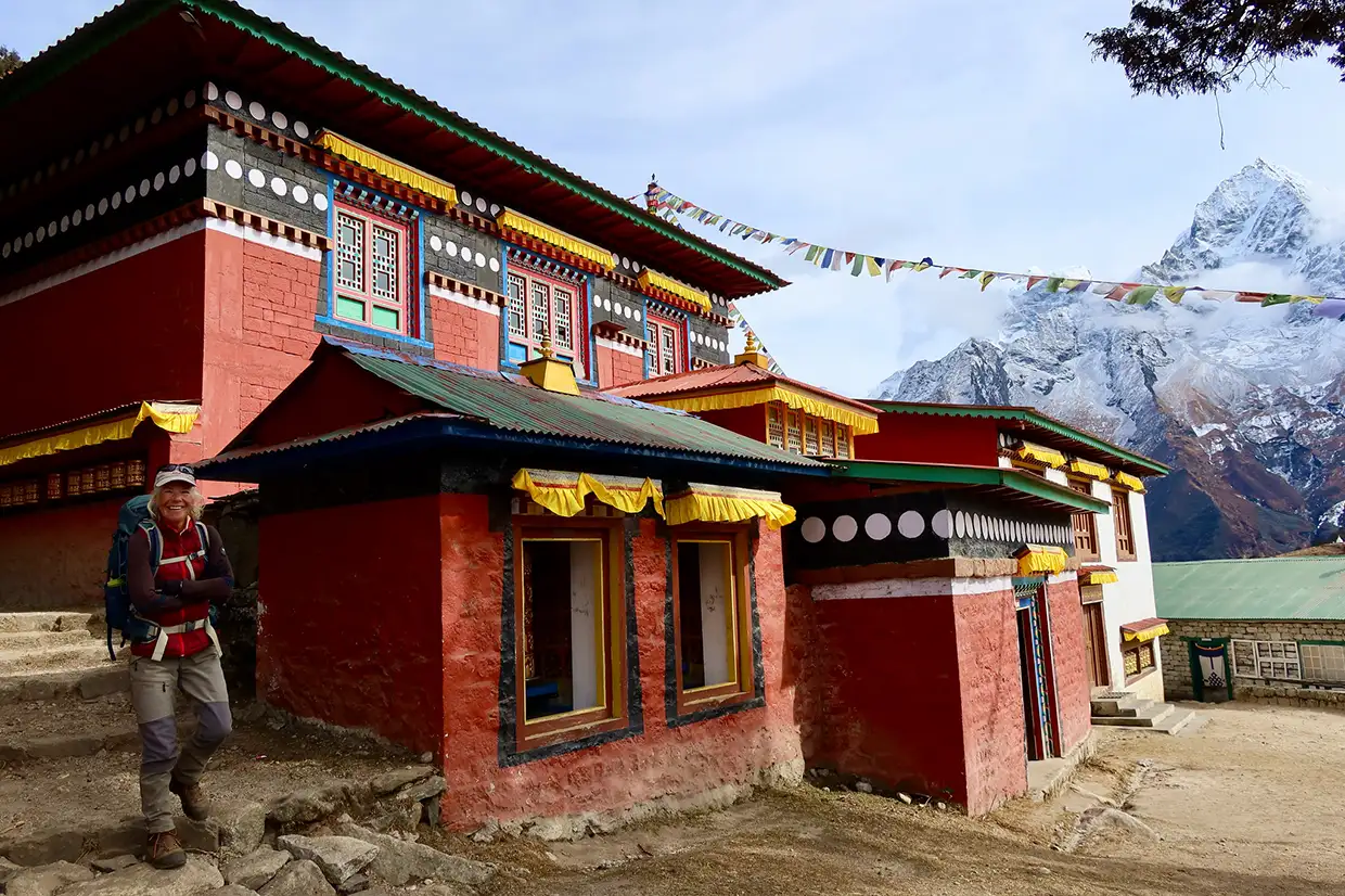 A trekker stands near the colorful Khumjung Monastery with prayer flags and snow-covered peaks visible during an Everest Cultural Trek in Nepal.