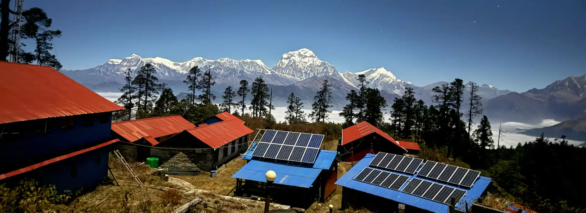 A detailed wide-angle landscape photograph looking over a rustic village with red and blue-roofed buildings towards the massive, detailed Himalayan peaks of Dhaulagiri and Nilgiri in the distance during the Mohare Danda Trek.