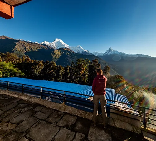 A trekker stands on a stone terrace looking out at the massive Annapurna and Machapuchare mountain peaks during the Khopra Ridge Trek.