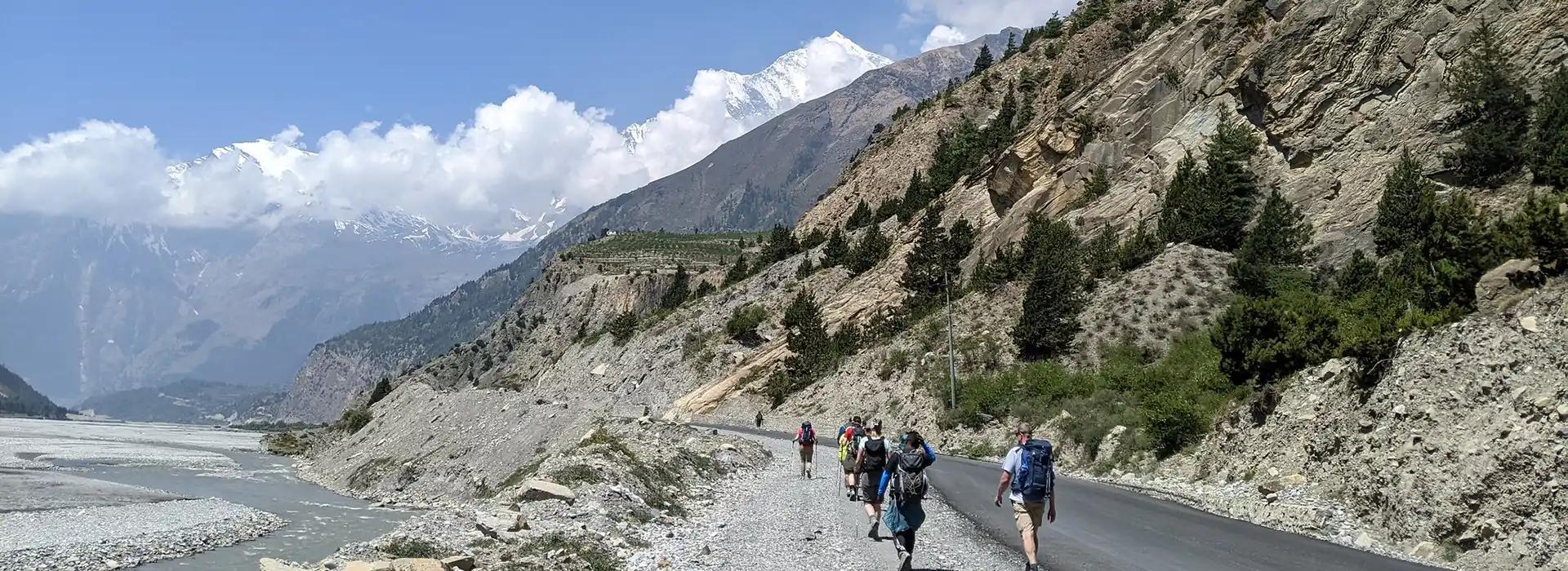 A group of hikers with backpacks walks along a winding mountain road next to a river toward snow-capped peaks, following their Annapurna Base Camp Trek Packing List.