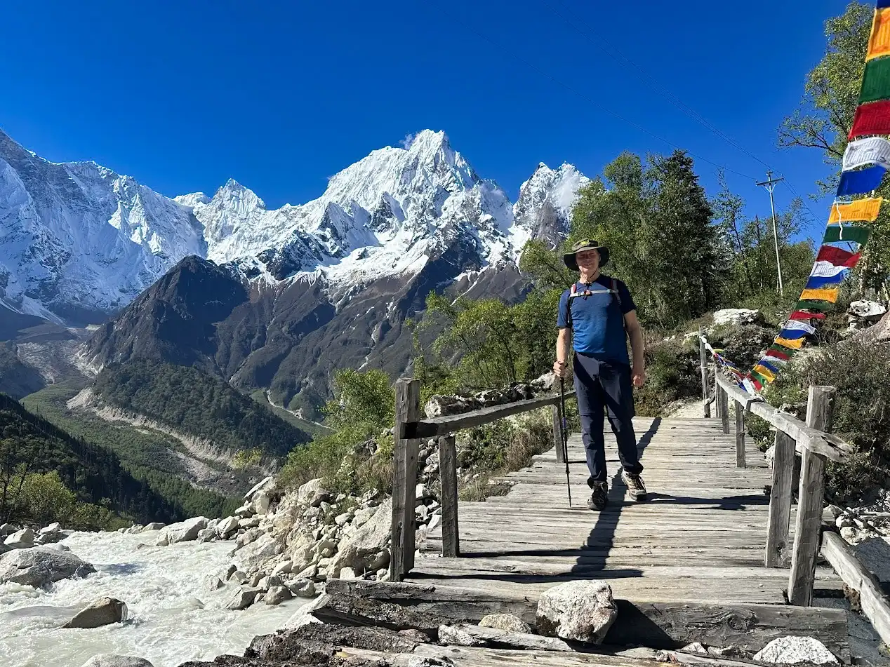 A traveler crosses a rustic wooden bridge over a rushing mountain river, with snow-capped Himalayan peaks rising sharply under a clear blue sky, featured in our Manaslu Circuit Trek Guide.