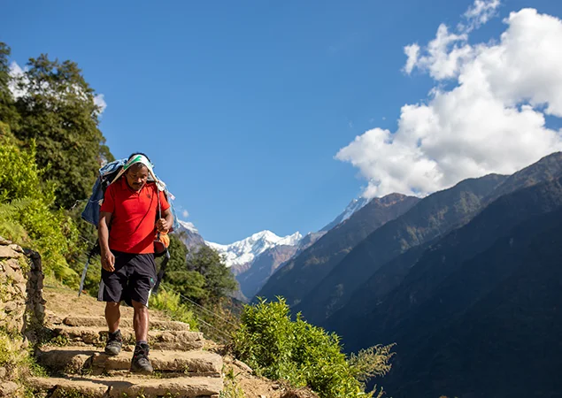A local porter descends steep stone stairs with a heavy pack against a backdrop of snow-capped peaks, demonstrating the Annapurna Base Camp Trek Difficulty.