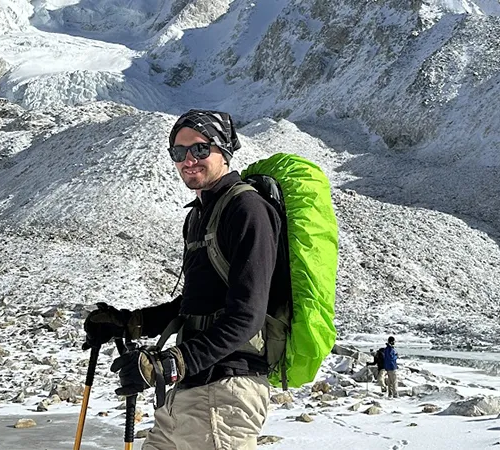 A hiker wearing sunglasses and a backpack with a bright green rain cover stands on a snowy glacial moraine trail, ideal for calculating the Manaslu Circuit Trek Cost.