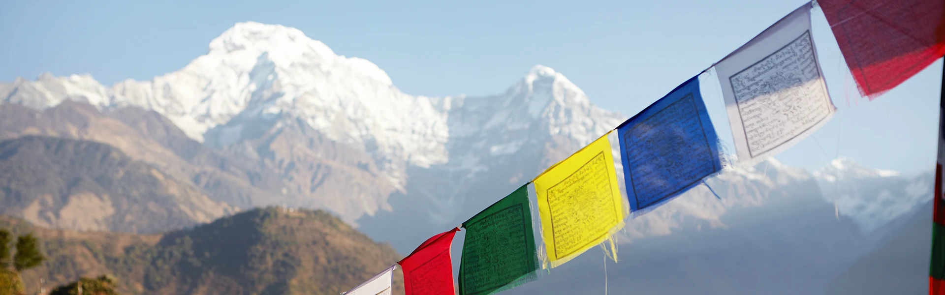 Colorful Buddhist prayer flags stretch across the foreground with massive, snow-covered Himalayan peaks in the background, illustrating a serene moment despite the Annapurna Base Camp Trek Difficulty.