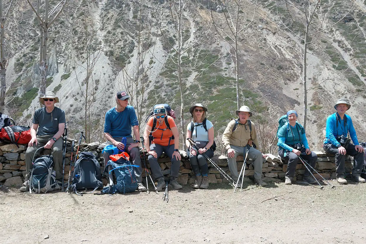A group of seven trekkers sits on a low stone wall resting with their backpacks and hiking poles during the Nar Phu Valley with Annapurna Circuit Trek.