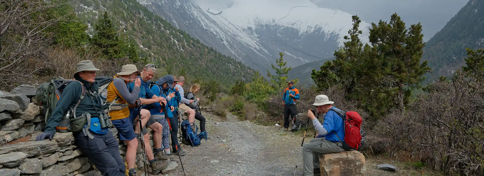 A group of hikers rests along a dirt mountain trail on a stone wall, facing a massive snow-covered peak during the Nar Phu Valley with Annapurna Circuit Trek.